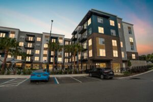 Modern apartment building with sunset reflections, parking area, and palm trees in the foreground.