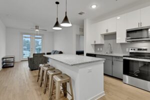 Modern kitchen with island, bar stools, stainless appliances, and open living area, featuring wood flooring and stylish lighting.