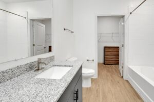 Modern bathroom with granite countertop, white tiles, wood flooring, and attached walk-in closet. Clean and bright design.