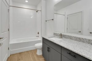 Modern bathroom with granite countertop, white tiles, wooden floor, bathtub, and large mirror reflecting light.