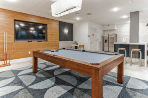 Modern game room with pool table, flat-screen TV, and kitchen bar area, featuring contemporary decor and seating.