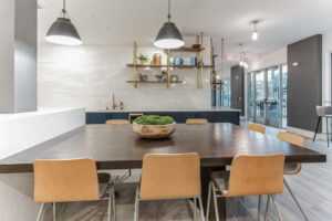 Modern communal dining area with wooden table, chairs, pendant lights, and decorative shelves in a bright interior space.