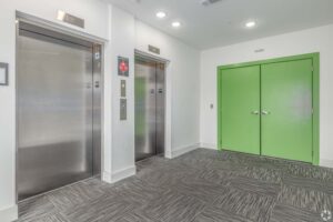 Two shiny elevators and a bright green door in a modern, well-lit hallway, featuring gray patterned carpet.