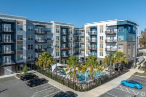 Modern apartment complex with palm trees and pool, surrounded by parked cars on a sunny day.