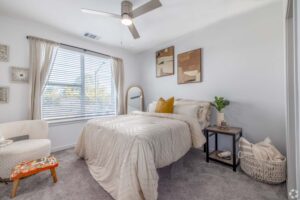 Stylish bedroom with cozy bedding, decorative pillows, wall art, and natural light from a large window.