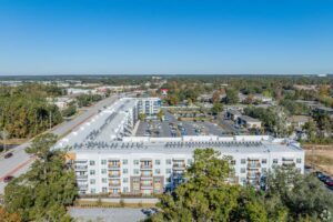 Aerial view of a modern apartment complex with parking, surrounded by trees and roads under a clear blue sky.