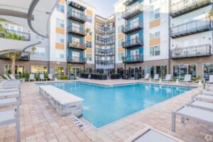 Modern apartment complex with a pool, lounge chairs, and balconies under a clear sky.