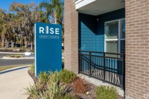 Sign reading Rise Thirty Fourth in front of a blue building with landscaped greenery and trees in the background.