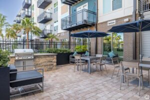 Modern apartment patio with outdoor seating, BBQ grill, and umbrellas surrounded by palm trees and stylish building.