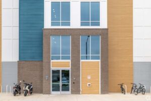 Modern building entrance with large windows, brick facade, and parked bicycles and scooters in front.